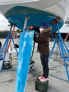 The rudder and post were dropped out of the boat and the rudder seals inspected. Here, Charity Hosler, one of many friends who helped get Twelve-Winded Sky ready for the race, sands the rudder.